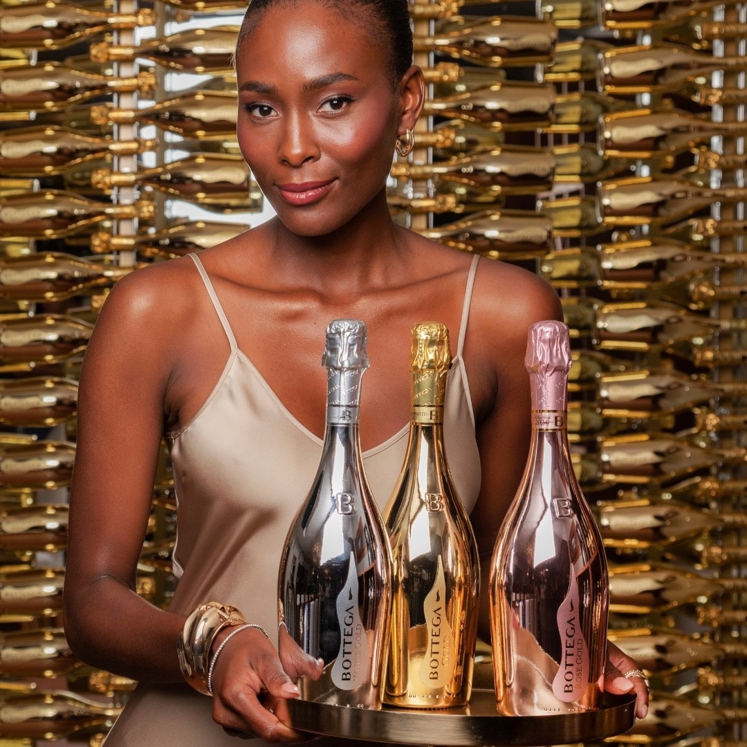 Woman in a beige slip dress presents a tray with three Bottega sparkling wine bottles (silver, gold, and rose) against a wall of wine racks.