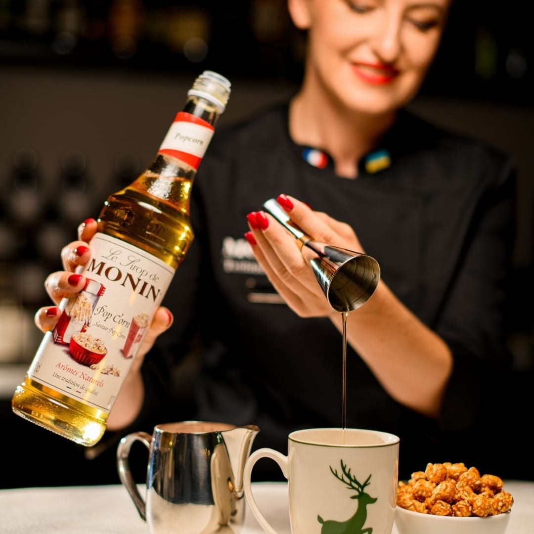 Bartender pours MONIN popcorn syrup into a mug with a green deer logo, hands with red nail polish visible, a metal creamer nearby and a bowl of caramel nuts on the counter.