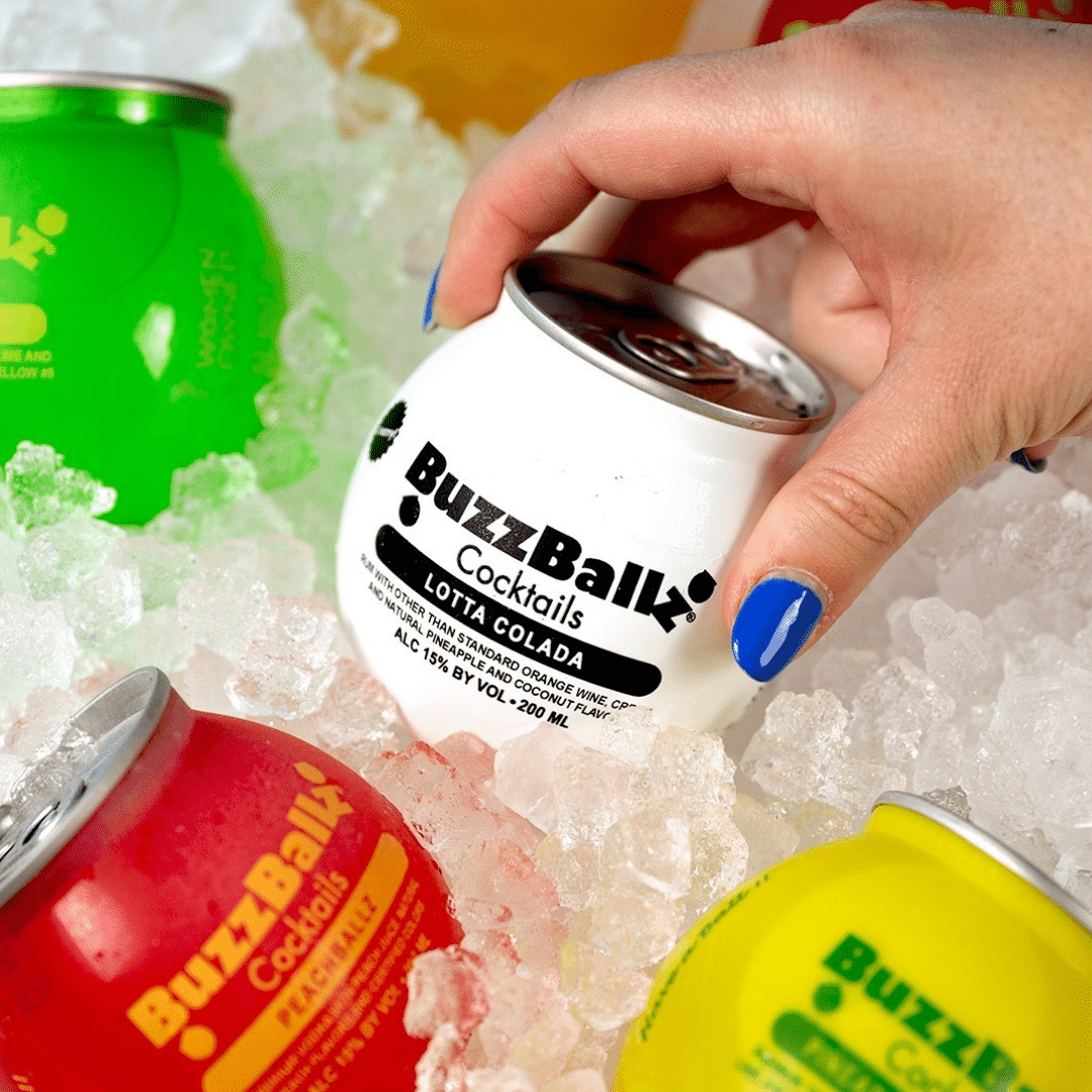 Person opening a white BuzzBallz cocktail can (Lotta Colada) among colorful cans on crushed ice.