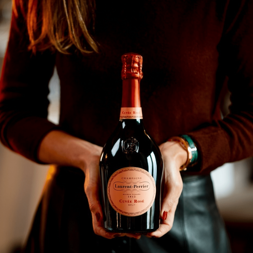 Person in a burgundy sweater holding a Laurent-Perrier Cuvee Rosé champagne bottle toward the camera.