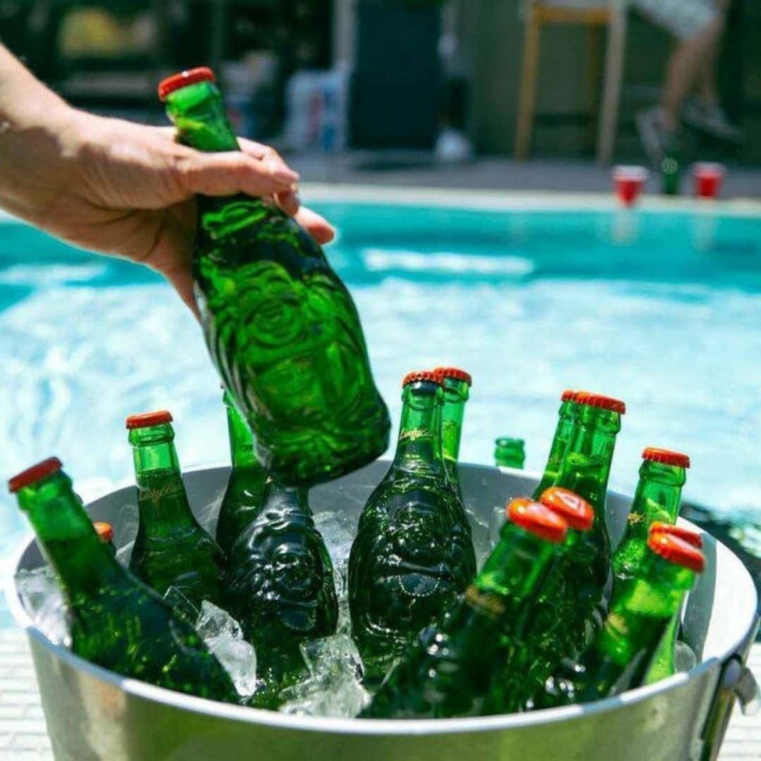 Person grabbing a green glass bottle from a ice-filled bucket by a poolside party scene.