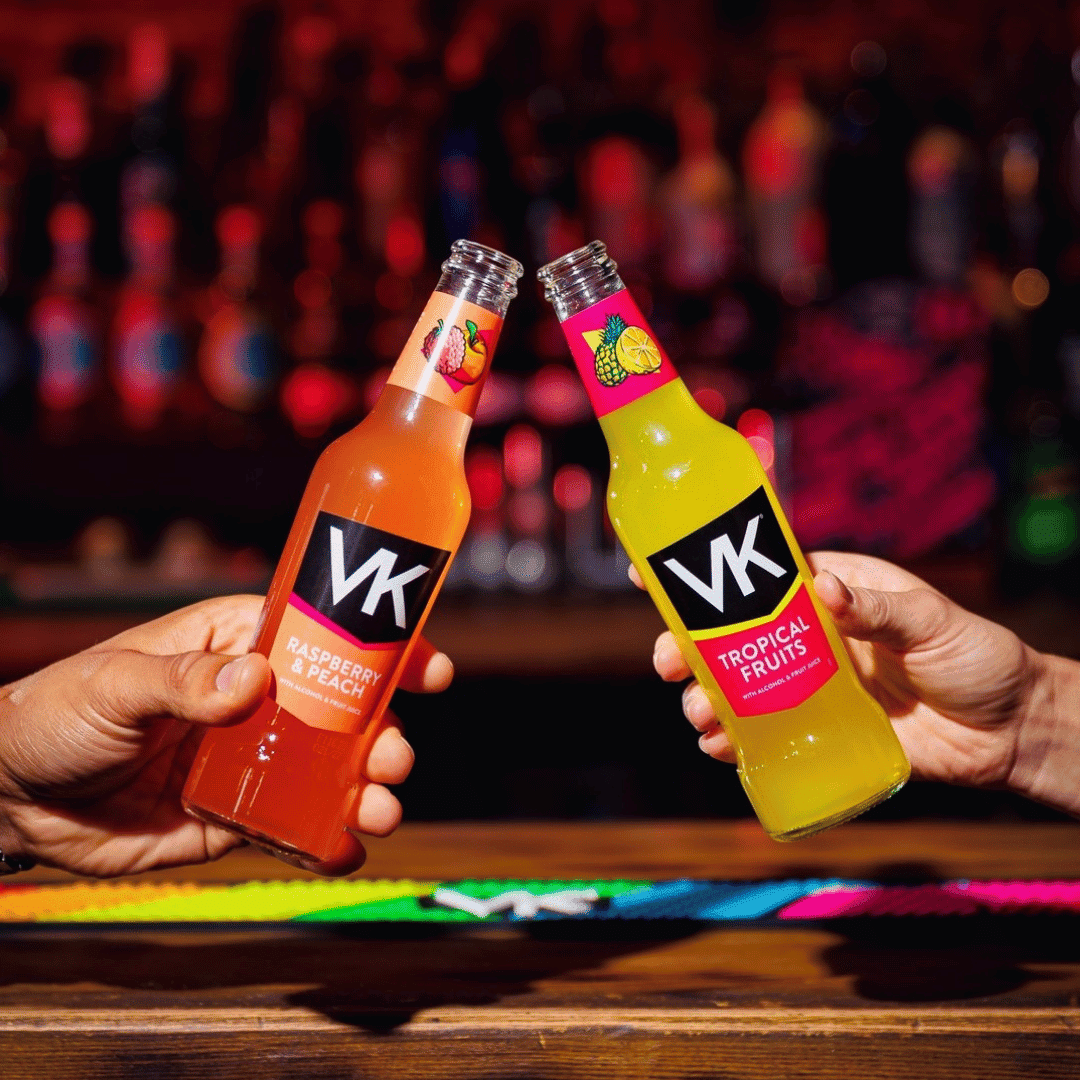 Two hands clink VK fruit-flavored bottles—raspberry & peach and tropical fruits—over a bar counter.