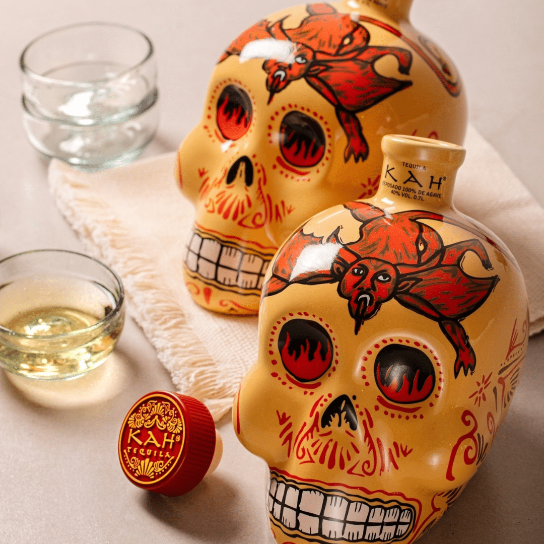 Two yellow Day of the Dead skull tequila bottles with red and black designs, cap nearby, and a shot glass on the side.