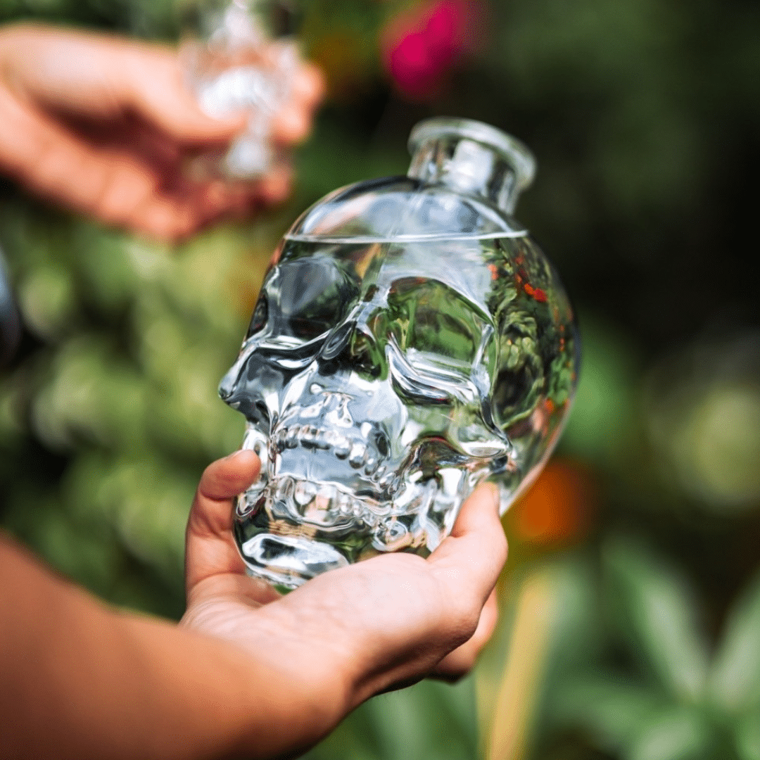 Hands hold a clear glass skull-shaped bottle outdoors, reflecting greenery in the glass